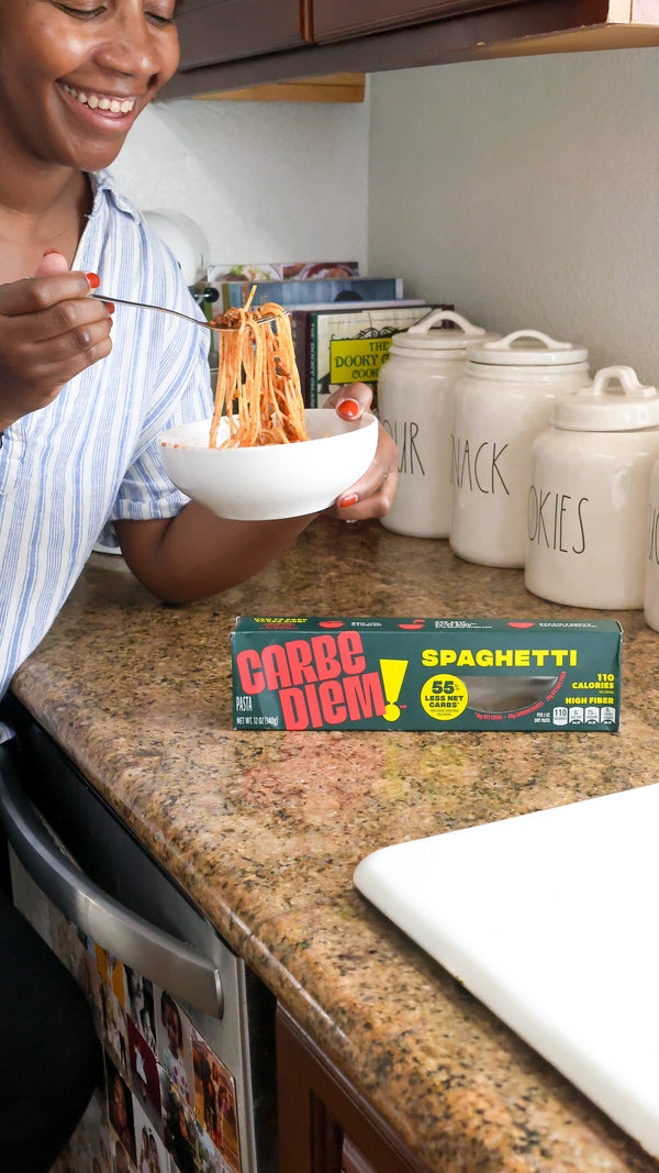 Person eating spaghetti from a bowl with a box of Carbe Diem Spaghetti on the counter.