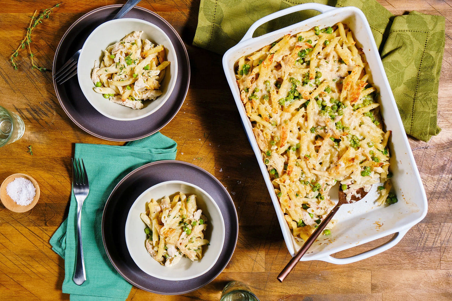 Baked pasta dish with peas served on a wooden table with plates and a green napkin.