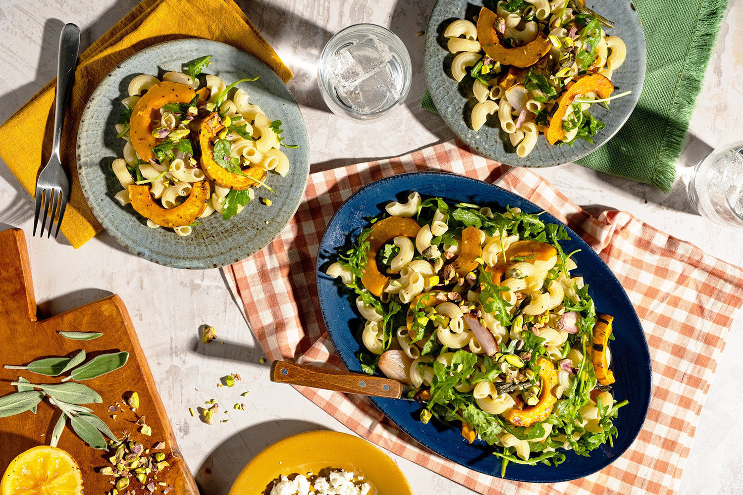 Three plates of pasta salad on a checkered tablecloth with utensils and ingredients.