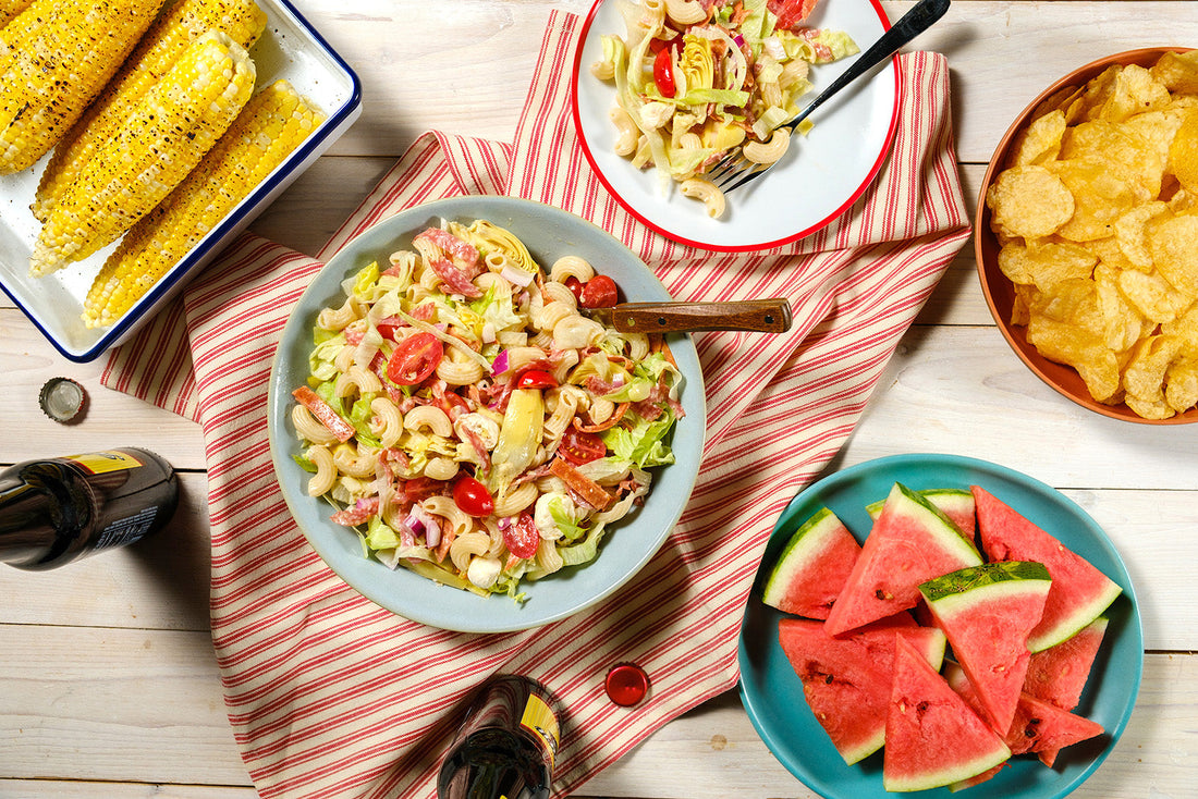 Outdoor setting with pasta salad, corn on the cob, watermelon, and chips on a wooden table.