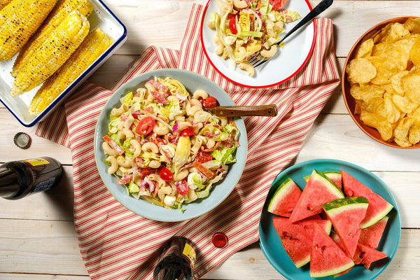 Outdoor setting with pasta salad, corn on the cob, watermelon, and chips on a wooden table.