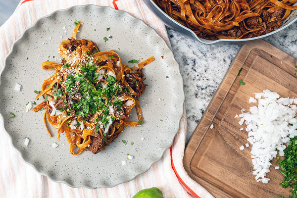 Plate of pasta with toppings on a textured surface with a cutting board and ingredients.