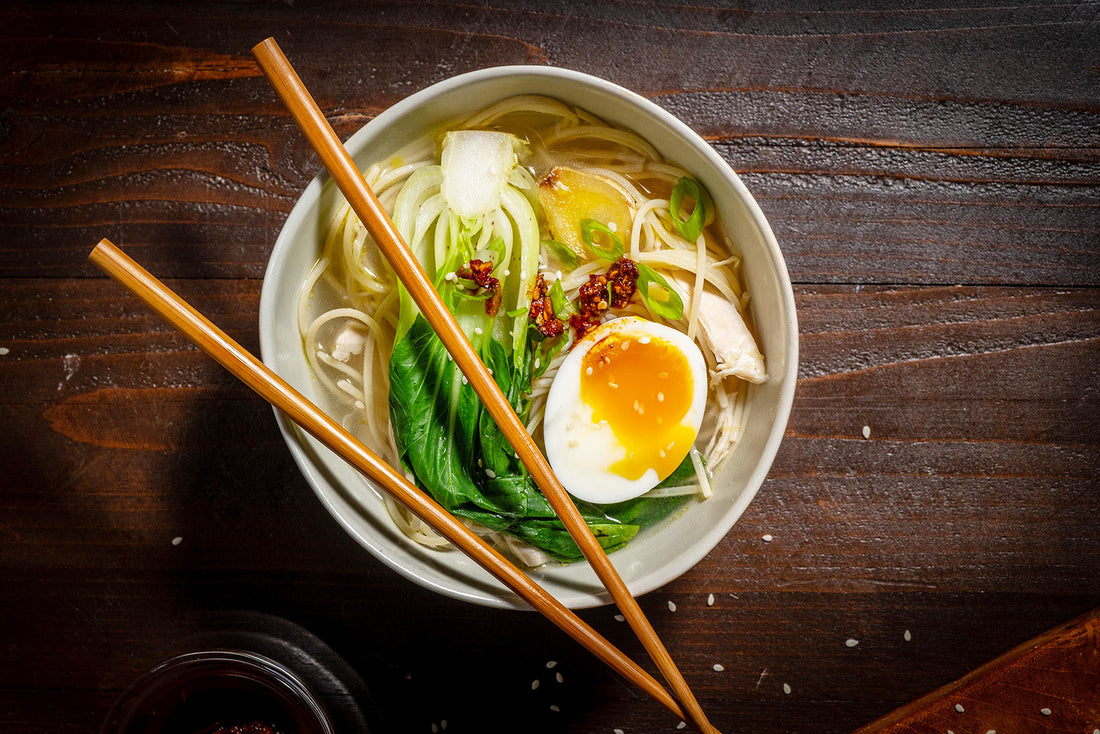 Bowl of noodles with a soft-boiled egg, green leaves, and sesame seeds on a wooden surface.