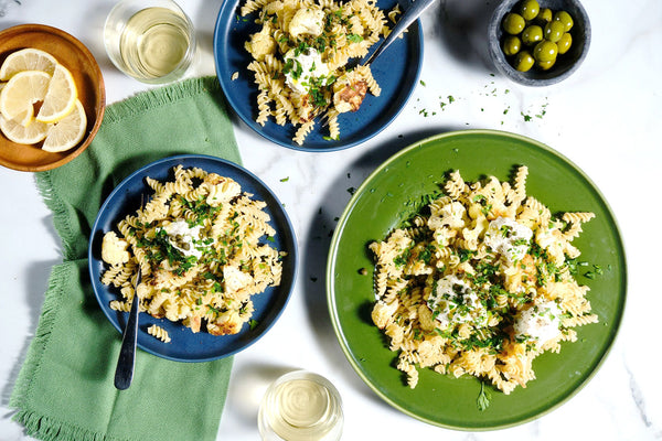 Plates of pasta with herbs and cheese, accompanied by glasses of white wine and a bowl of olives on a white tablecloth.