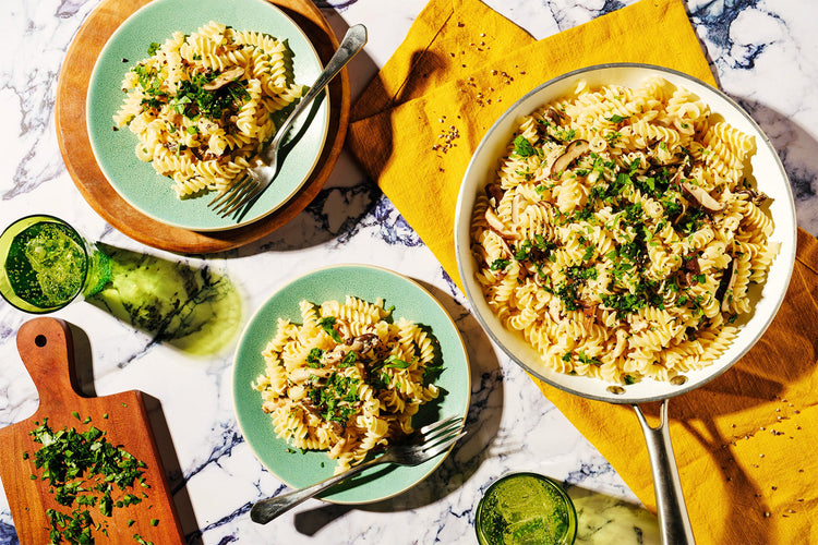 Pasta dish with greens served on a marble table with a wooden cutting board.