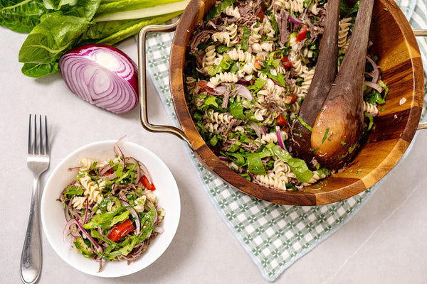 Pasta salad in a wooden bowl with a side dish and ingredients on a checkered tablecloth.