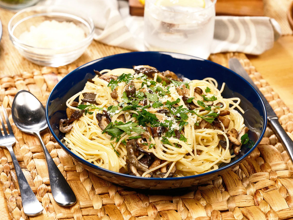 Bowl of spaghetti with mushrooms and herbs on a woven placemat with cutlery.