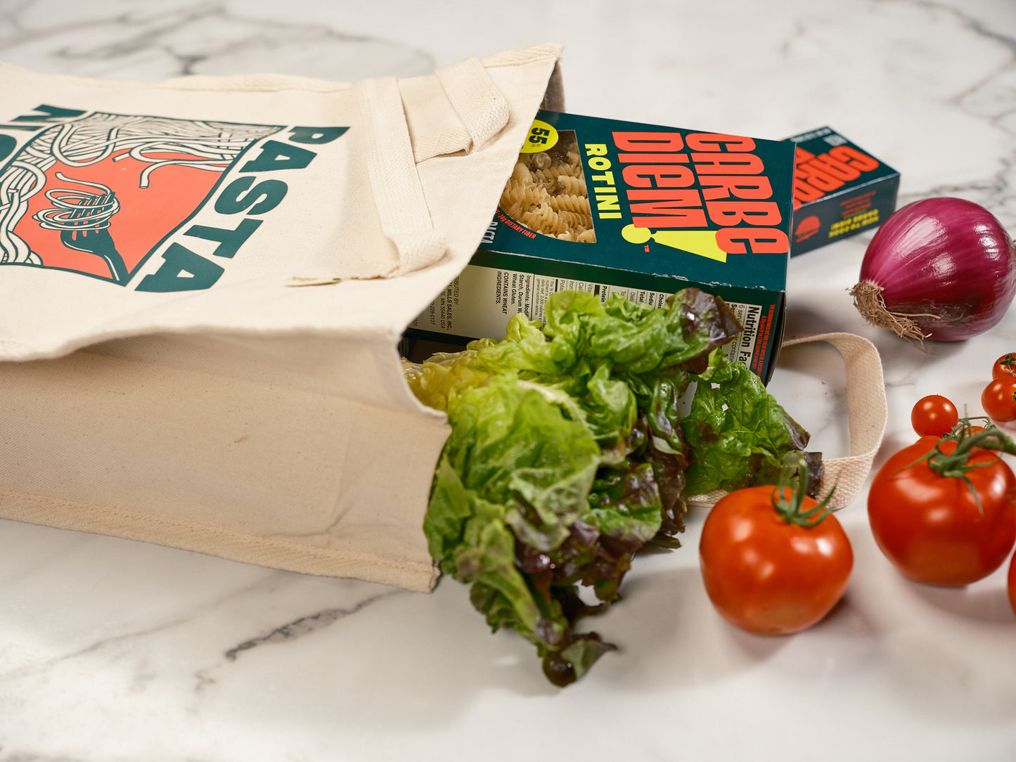 Cream-colored canvas tote bag with vegetables spilling out. Tote bag has an mage of spaghetti and reads "Pasta Night"