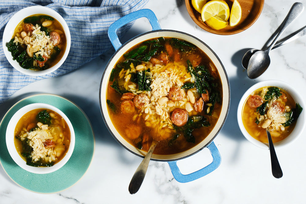 Pasta soup with greens and sausage in a blue pot, surrounded by bowls of soup on a marble surface.