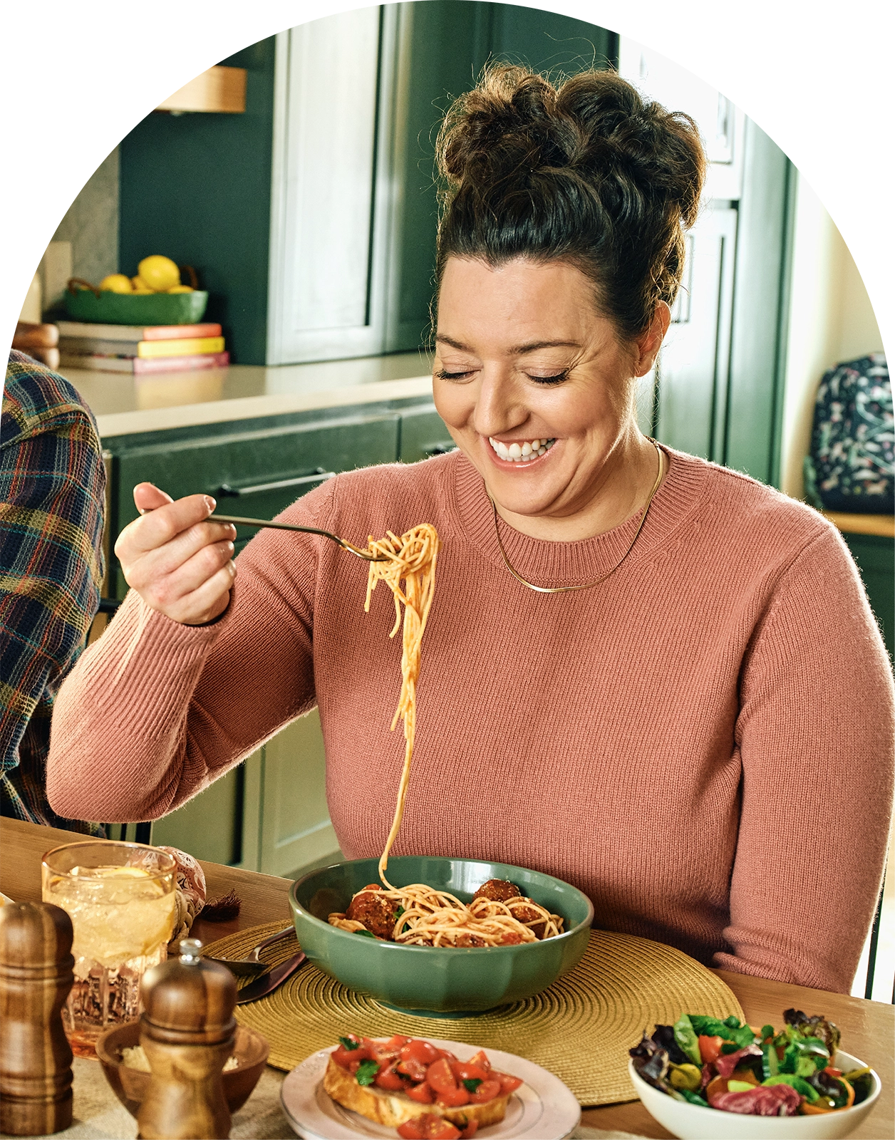 Woman eating spaghetti in a kitchen with a bowl of salad and bread on the table.
