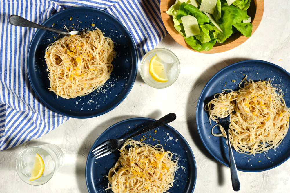 Three blue plates with spaghetti, a bowl of salad, and glasses of water on a white tablecloth.