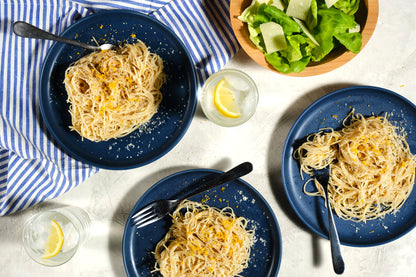 Three blue plates with spaghetti, a bowl of salad, and glasses of water on a white tablecloth.