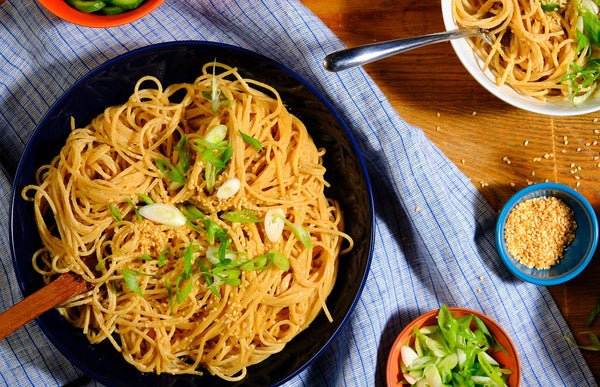 Noodles with green onions in a black bowl on a wooden table with a blue checkered cloth.