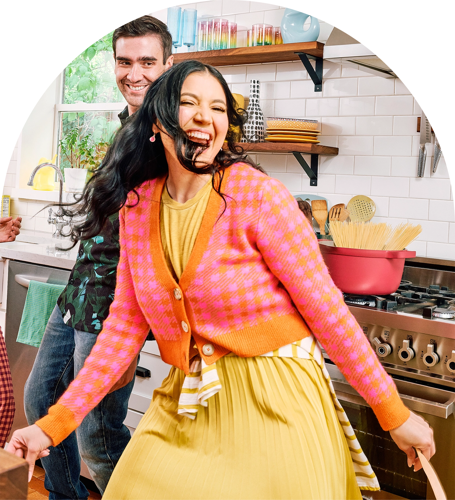 Two people in a kitchen, one wearing a colorful pink and orange cardigan and yellow skirt, the other in a green shirt.