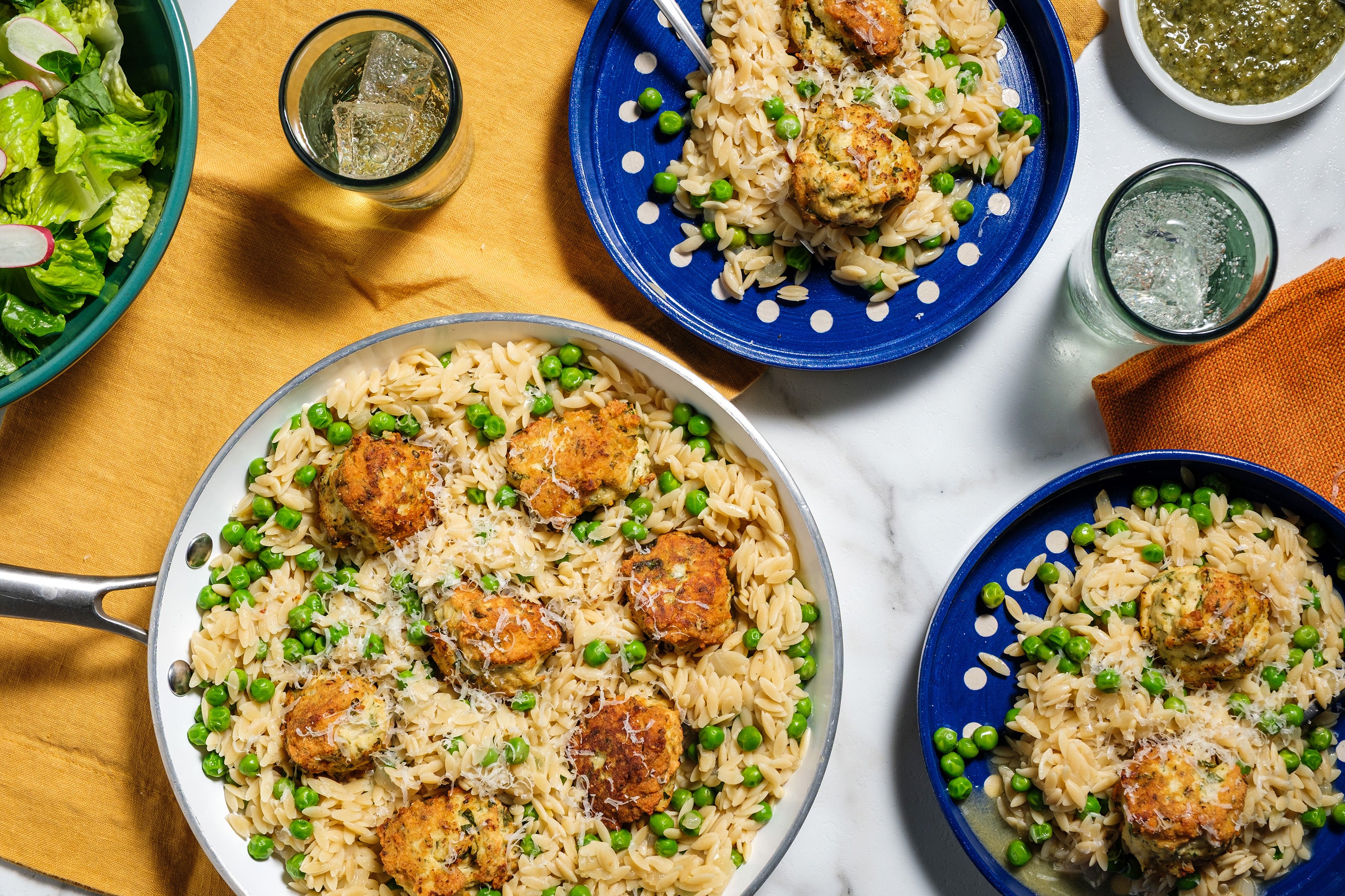 Orzo with chicken meatballs and peas served in a pan and on plates, with drinks on the side.