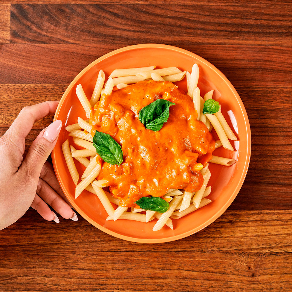 Pasta dish with tomato sauce and basil leaves on an orange plate, held by a hand on a wooden table.